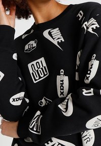 Svart sweatshirt med vita tryckta logotyper, inklusive Nike och Air Max, med rund halsringning och ribbade ärmslut. Mjuk bomullsblandning genom texturen.