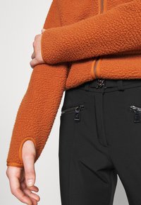 Gros plan d'une personne portant une veste polaire orange texturée avec un trou pour le pouce et un pantalon noir avec des poches avant zippées.