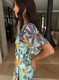 Vestido colorido com padrão em azul claro, com desenhos florais e gráficos. Apresenta mangas curtas e fluídas e uma cintura ajustada. Brincos dourados complementam o visual.