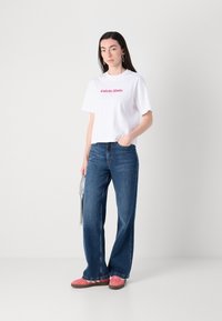 Balts īss T-krekls ar sarkano "Calvin Klein" tekstu, apvienots ar zilām platajām biksēm un rozā kedām. Modelis tur sudraba soma.