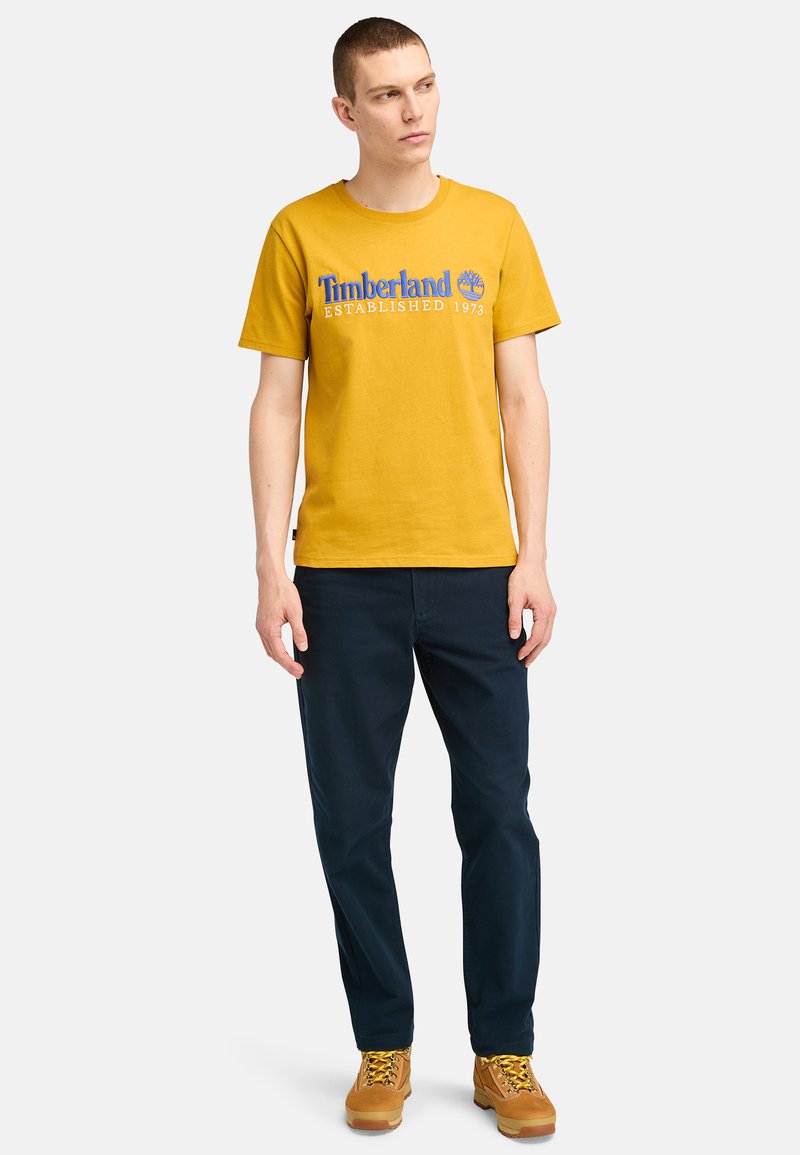 Żółta bawełniana koszulka z niebieskim logo "Timberland" i datą założenia. Połączona z granatowymi spodniami i brązowymi butami z żółtymi sznurówkami.