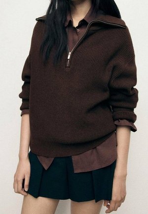 Femme portant un pull en tricot marron foncé à fermeture éclair par-dessus une chemise marron à col et une jupe plissée noire, debout devant un fond uni.