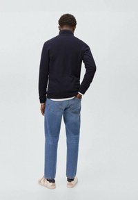 Mornarsko pleteno pulover s visokim ovratnikom, v kombinaciji s svetlo modrimi hlačami ravnega kroja in bež supergami, prikazan od zadaj.