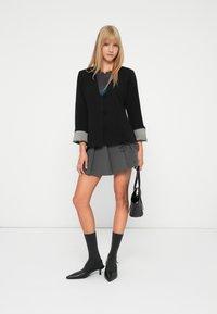 Jonge vrouw met blond haar, gekleed in een zwarte blazer, een grijze geplooide rok, donkere sokken, zwarte hakken en een zwarte handtas, tegen een witte achtergrond.