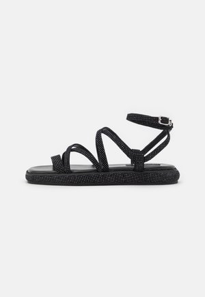 Schwarze flache Sandalen mit mehreren überkreuzten Riemen; das Material weist kleine weiße Punkte auf; Knöchelriemen mit Schnalle und kontrastierender strukturierter Sohle.