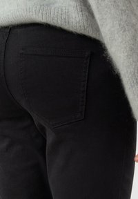 Detailní záběr osoby v černých džínách, z nichž je vidět zadní kapsa, a šedém svetru, který částečně zakrývá oblast pasu.