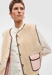 Μπεζ φλις γιλέκο με καφέ περιθώριο, διακοσμημένο με χρυσά κουμπιά και υφασμένη ροζ τσέπη. Φορεμένο πάνω από λευκό πουκάμισο με σχέδια.