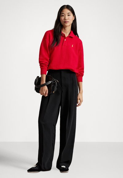 Rød langermet poloshirt med krage, svarte vide bukser og en svart skulderveske. Modellen har på seg svarte tøfler.