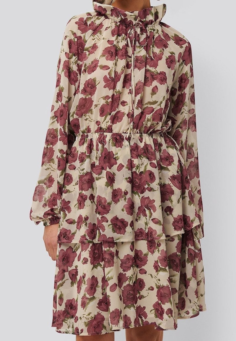 Robe florale avec un fond crème, ornée de roses bordeaux, à manches longues, col volanté et taille élastique avec une jupe superposée.