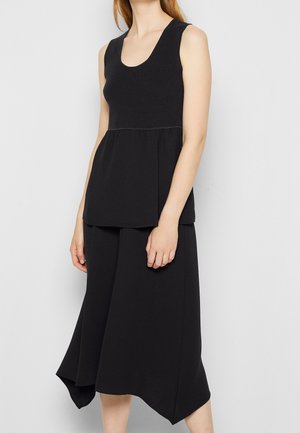 Femme portant un haut noir sans manches à texture côtelée et une jupe noire asymétrique sur un fond uni clair.