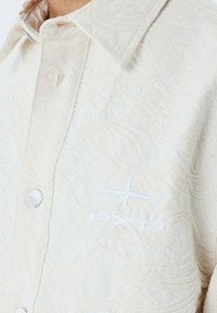 Crèmewitte shirt met een gestructureerd patroon, voorzien van een knoopsluiting aan de voorkant en een geborduurd logo op de linkerborst.