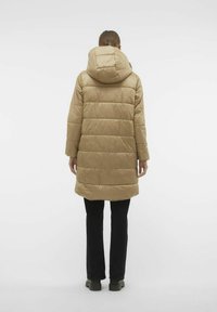 Beige pufferjas met een capuchon, gekenmerkt door een gestikt ontwerp en een middellange snit. Gepaard met zwarte broek en groene laarzen.