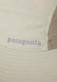 Bežs cepure, izgatavota no auduma, ar izliekto malu. Satur kontrastējošu brūnu paneļu un pelēku "Patagonia" logo. Redzami šuvju detalizējumi.