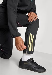 Sorte treningsbukser med gule tre-stripe design, rosa sømmer og glidelåsdetaljer ved ankelen. Kombinert med svarte fotballsko og hvite sokker.