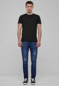 Camiseta negra ajustada, vaqueros ajustados azules desgastados con detalles rasgados y zapatillas en blanco y negro. El modelo está de pie contra un fondo gris.