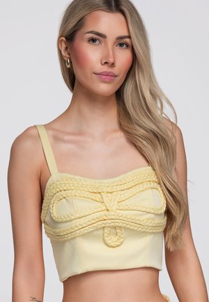 Jeune femme aux longs cheveux blonds portant un crop top jaune clair avec des détails en corde tressée, debout devant un fond uni.