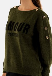 Maglione verde oliva realizzato in materiale morbido e peloso. Presenta un collo a girocollo, testo nero audace e bottoni decorativi argentati sulle spalle.