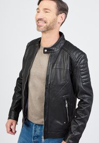 Sort læderjakke med høj krave, lynlås foran, kviltede skulderdetaljer og sidelommer. Bruges over en lys grå top.