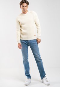 Suéter de punto acanalado color beige claro con cuello redondo, combinado con jeans azules y zapatillas blancas. Presenta un pequeño detalle del logo en el suéter.