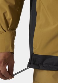 Veste imperméable de couleur brun doré avec des emmanchures noires, dotée de cordons réglables à l'ourlet et d'une finition en tissu texturé.