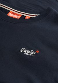 Camiseta de algodón azul marino con cuello redondo, logotipo bordado de "Super Dry" en blanco y naranja, con cuello acanalado y textura sutil.