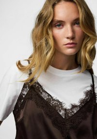 Brun sateng kjole med blondekanter lagd over en hvit t-skjorte. Håret er bølget og rammer inn ansiktet, med en nøytral bakgrunn.