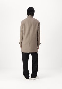 Un abrigo de lana marrón con corte recto, cuello alto y una abertura en la parte trasera, combinado con pantalones negros y zapatillas blancas.