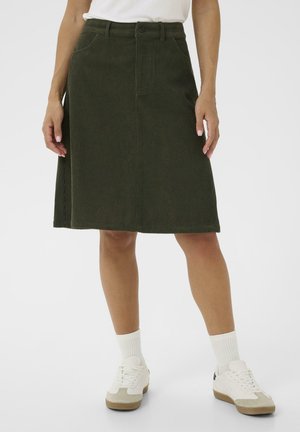 Zielona spódnica sztruksowa w kształcie litery A z talią o średnim stanie, zapinana z przodu na guziki, z szlufkami na pasek, zestawiona z białymi tenisówkami i prążkowanymi skarpetkami.