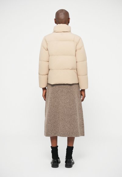 Beige puffer jacket with a high collar, quilted design, and long sleeves, paired with a brown textured skirt and black ankle boots.