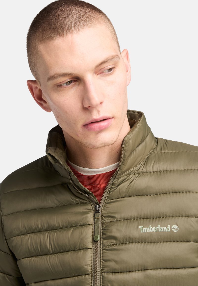 Oliwkowa, puchowa kurtka z zamkiem z przodu, stójką i delikatnym logo. Wykonana z gładkiego, izolowanego materiału z poziomym przeszyciem.