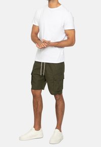 Homme portant un T-shirt blanc uni, un short cargo vert olive avec un cordon de serrage, et des baskets blanches, debout les mains jointes.