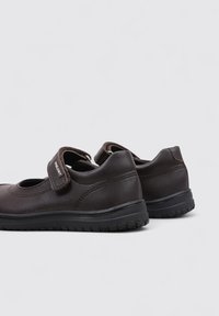 Zapatos de cuero marrón con punta redondeada, correa segura y suela de goma texturizada, que presentan sutiles detalles de costura y un diseño acolchado.