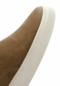 Scarpa slip-on in suede marrone con design curvo, suola in gomma bianca e cuciture visibili lungo il bordo. Texture liscia con dettagli minimi.