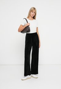 Jonge vrouw in een wit t-shirt, zwarte wijde broek, witte sneakers, terwijl ze een bruine schoudertas vasthoudt tegen een effen lichte achtergrond.