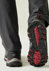 Persona con pantalones negros caminando, mostrando la suela con dibujo en negro y rojo de un zapato de senderismo sobre un fondo claro.