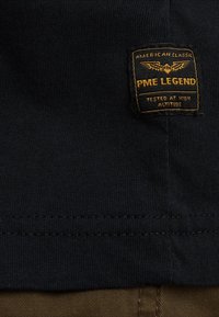 Zwarte katoenen T-shirt met textuur; heeft een vierkant geweven label in oranje en geel met "PME LEGEND" en vleugelsontwerp.