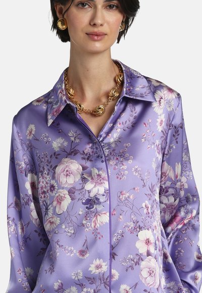Woman wearing a purple satin floral blouse with a pointed collar, paired with gold statement earrings and a chunky gold chain necklace.