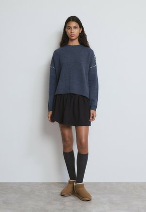 Woman wearing a blue sweater with pearl details, black skirt, knee-high dark socks, and tan shearling boots standing against a plain wall.