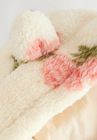 Tissu en polaire couleur crème avec une texture douce, présentant des motifs abstraits rose et vert. Comprend un détail en pompon duveteux.
