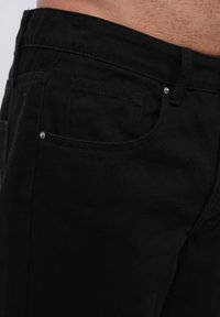 Jeans de mezclilla negra, con un diseño ajustado, bolsillos delanteros y herrajes en tono plateado con una superficie de tela ligeramente texturada.