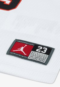 Valge võrgukangast plaastriga, millel on mustad ja punased aktsendid, kujutades Jumpman logo ja numbrit 23. Tekstiks on "Brand of Flight."