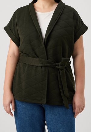 Person trägt eine kurzärmelige, gesteppte dunkelgrüne Wickeljacke mit Stoffgürtel, der in der Taille gebunden ist, über einem weißen Hemd und blauen Jeans.