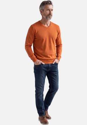 Uomo con capelli e barba grigi che indossa un maglione arancione a scollo a V, jeans blu scuro e scarpe di pelle marroni, in piedi con le mani in tasca.