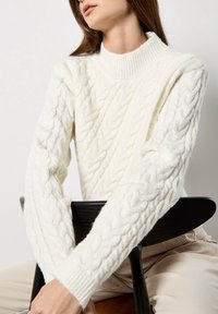 Maglione bianco a trecce con collo alto a coste e maniche lunghe, caratterizzato da un motivo testurizzato e un tessuto morbido. Modello seduto su una sedia.