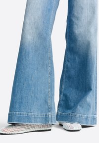 Svetlomodré široké rifle s vypranou textúrou a otrhanými lemami, doplnené otvorenými sieťkovanými bielymi topánkami s čiernou podrážkou.