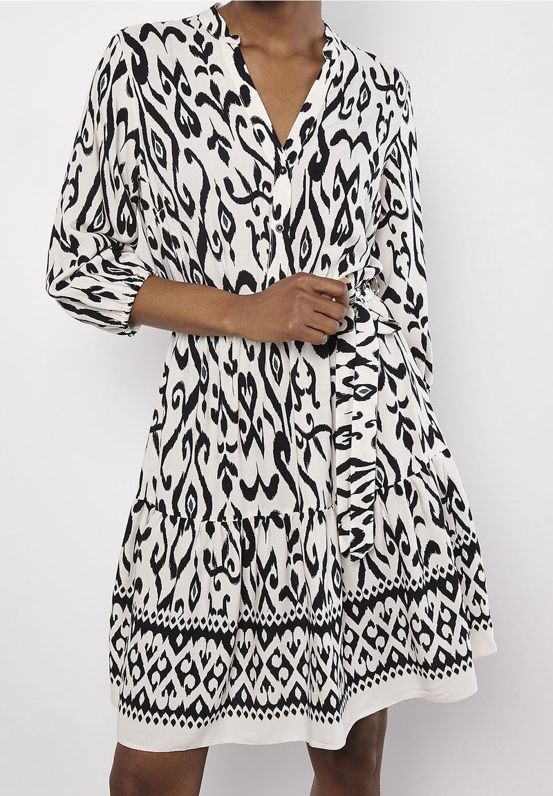 Femme portant une robe aux motifs noir et blanc, longueur genoux, manches 3/4, avec une ceinture nouée à la taille, sur un fond clair uni.