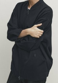 Henkilö, jolla on päällään tummansininen vetoketjullinen takki, jossa on nappikiinnitteiset taskut, kädet ristissä, seisoo vaaleaa taustaa vasten.