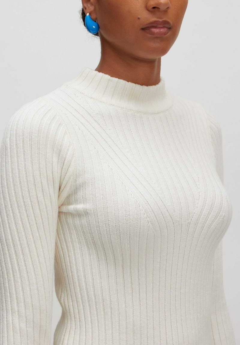 Lichtcrème geribde coltrui gemaakt van structuurstof. Beschikt over een aansluitende pasvorm en subtiele diagonale ribbeldetails.