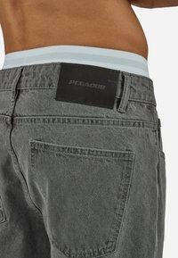 Man’s lower back wearing gray denim pants with a black leather brand patch labeled "PEGADOR" above the right back pocket.