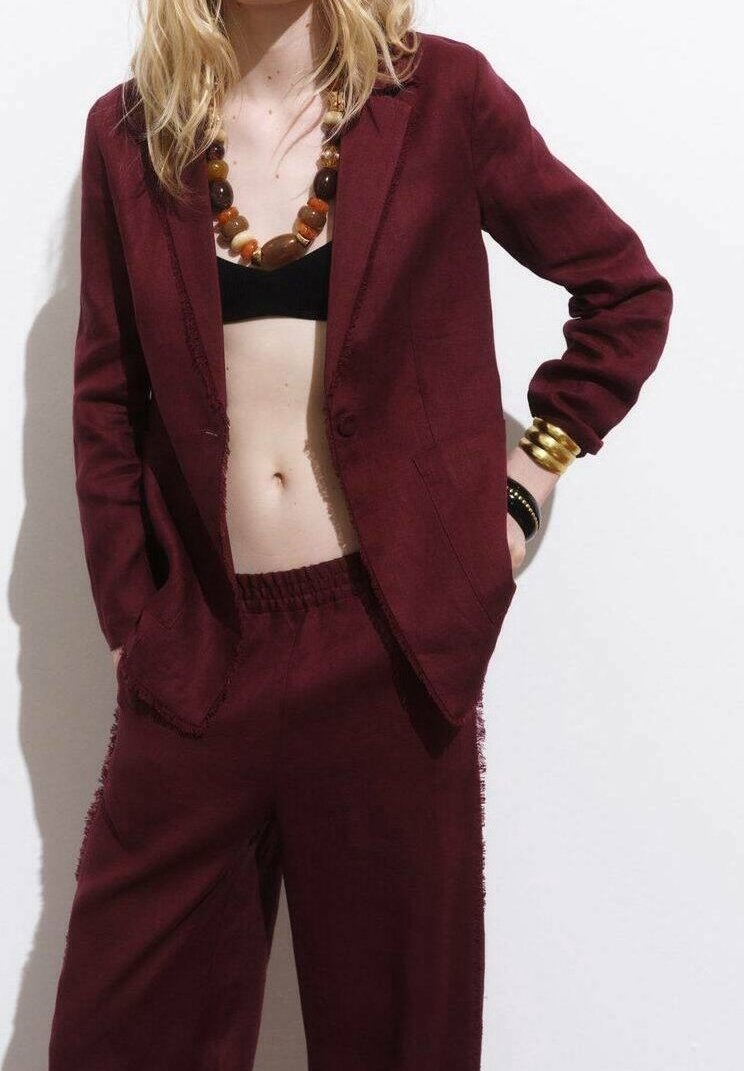 Femme portant un costume bordeaux avec blazer et pantalon, top court noir, collier en perles de bois et bracelets dorés, debout les mains dans les poches.